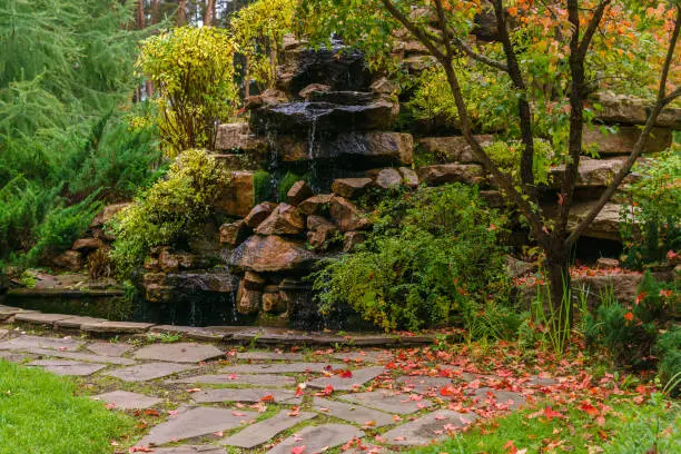 Photo of lawn among decorative bushes with a path and artificial rock with waterfall