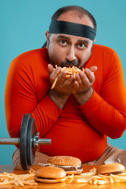 istock Middle-aged man with beard, dressed in a red turtleneck, headband, posing with burgers and french fries. Blue background. Close-up. Fast food 1174648954