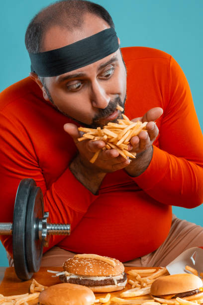 istock Middle-aged man with beard, dressed in a red turtleneck, headband, posing with burgers and french fries. Blue background. Close-up. Fast food 1174649014