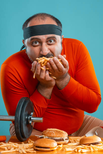 istock Middle-aged man with beard, dressed in a red turtleneck, headband, posing with burgers and french fries. Blue background. Close-up. Fast food 1174649186
