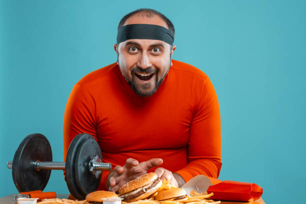 istock Middle-aged man with beard, dressed in a red turtleneck, headband, posing with burgers and french fries. Blue background. Close-up. Fast food 1174649889