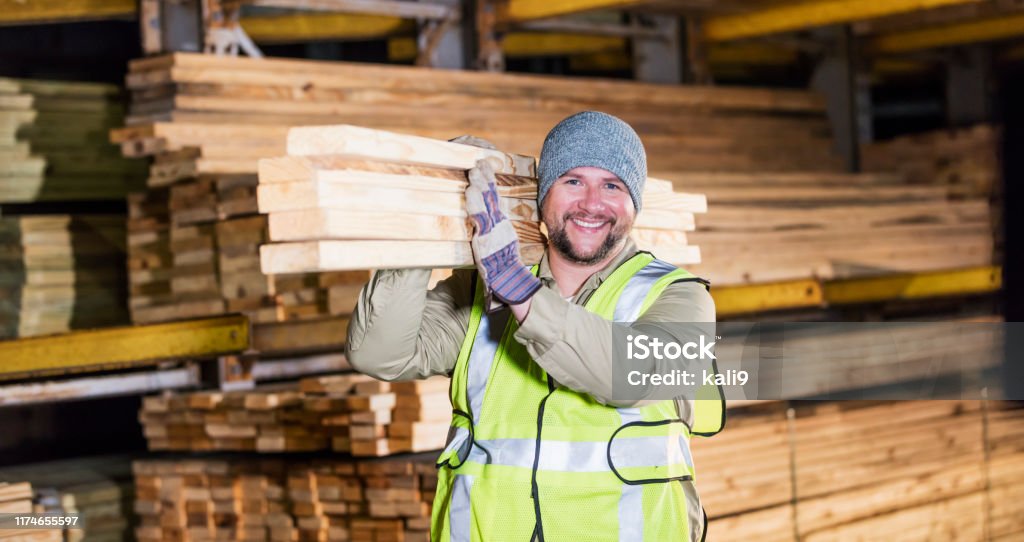 Hispanic man working at lumber yard A mid adult Hispanic man in his 30s working at a warehouse at a lumberyard or home improvement store. He is carrying a stack of lumber on his shoulder, smiling at the camera. Lumberyard Stock Photo