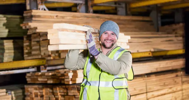 A mid adult Hispanic man in his 30s working at a warehouse at a lumberyard or home improvement store. He is carrying a stack of lumber on his shoulder, smiling at the camera.