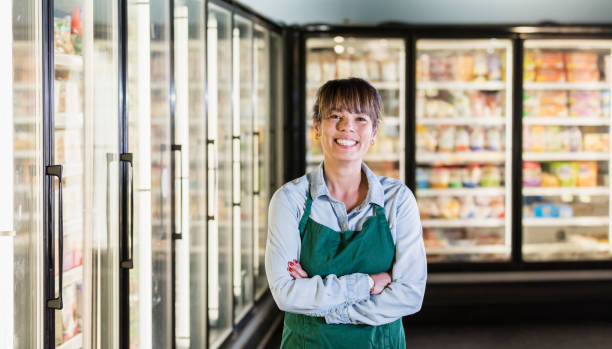 Employee in refrigerated section of supermarket A mature mixed race Asian and Caucasian woman working in a supermarket. She is standing with arms folded, in the frozen food or refrigerated aisle of the store, smiling at the camera. refrigerated section supermarket photos stock pictures, royalty-free photos & images