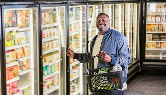 A mature African-American man in his 40s in a supermarket shopping for groceries.  He is in the frozen food aisle, about to open a refrigerator door, carrying a basket. He is smiling at the camera.
