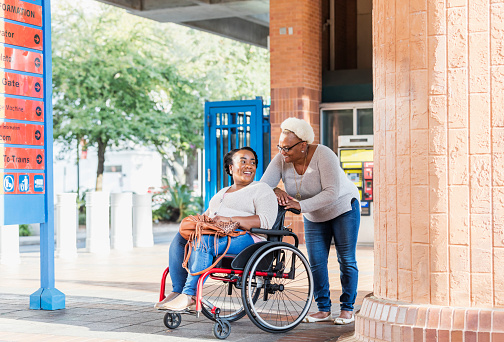 A mid adult African-American woman in her 30s in a wheelchair, outdoors in the city with her mother, a senior woman in her 60s, outside a train station. The daughter has spina bifida. They are smiling at the camera.