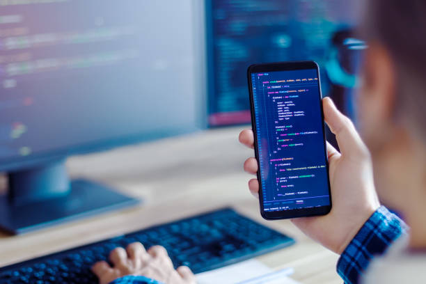 Software developer, freelancer working at home Closeup freelancer hand is holding smartphone with program code. Programmer developer is writing software on multiple computer screens at home office. Devices for working. Geek workplace concept. evolution stock pictures, royalty-free photos & images