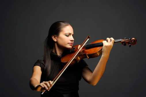 A young male is smiling and looking at a violin that he is holding up. He is wearing a black shirt and a vest sweater. He is in a studio with a isolated white background.