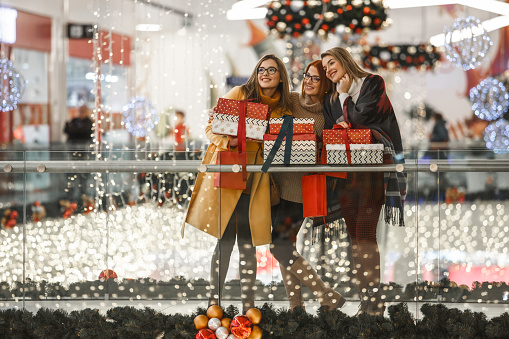 Copy space shot of three women posing with presents at the shopping mall. They are standing by the railing, surrounded by Christmas lights and decorations.