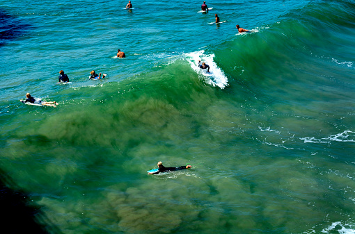Huntington Beach, CA / USA - Sept 7, 2019: Surfer rides a wave while his fellow surfers wait for the next wave. People watching the surfers from the Huntington Beach pier cast a shadow in the lower left hand corner.
