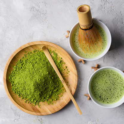 Matcha green tea ,powder matcha on a wooden round plate,welded powder in white Tawan,tasaku and a bamboo whisk on a light white concrete background