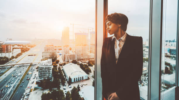 A businesswoman near the window A pensive African-American woman entrepreneur is thoughtfully looking at the cityscape outside the window while leaning on the window frame on the top floor of a luxury business office interior lean construction management stock pictures, royalty-free photos & images