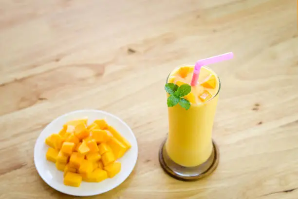 Mango smoothie in tall glass with plate of diced mangoes on wooden table.Beautifully peeled mango in white plate on wooden table.