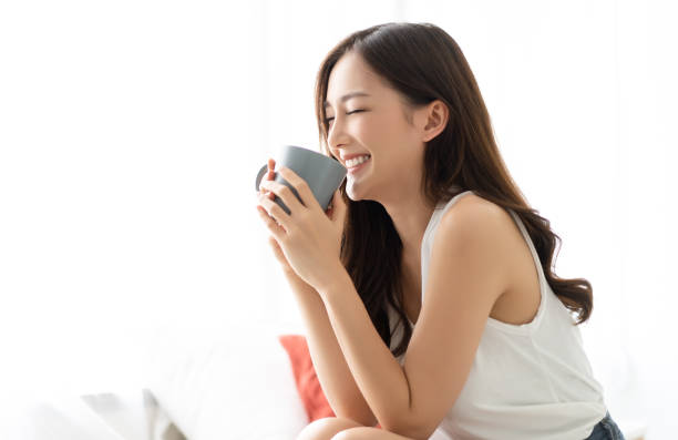 glücklich lächelnd schöne asiatische frau aufwachen mit tasse kaffee im schlafzimmer mit augen geschlossen. people lifestyle konzept; - chinese tea stock-fotos und bilder