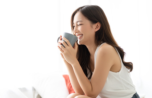 Happy smiling beautiful Asian woman awaking with cup of coffee at bedroom with Eyes Closed. People Lifestyle concept;