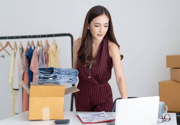 Young Asian casual woman working small business check her order in laptop computer and packing jeans to cardboard box in home office. Startup Small business owner,Online selling, e-commerce, delivery, freelance working concept. joven asiática mujer casual que trabaja pequeña empresa comprobar su pedido en computadora portátil y empacar jeans a caja de cartón en la oficina en casa. startup propietario de pequeñas empresas, venta en línea, comercio electrónico, entrega, conc