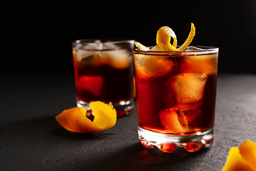 Negroni alcoholic cocktail in two glasses decorated with orange peel with ice cubes on a dark concrete table