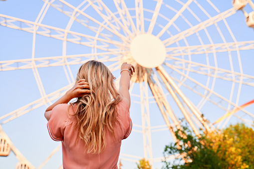 Back of stylish girl stands on street, wearing pink dress, shows her finger on wheel of review or ferris wheel. Beautiful horizontal shot of woman in an amusement Park, on background of carousel.