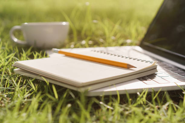 Notebook and pencil are placed on a laptop while the laptop and coffee cup on  green grass In the public park
