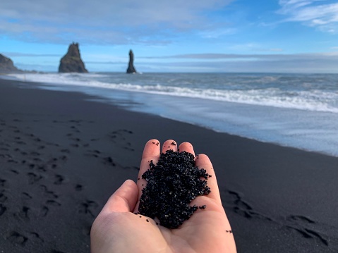 Black sand on the white female hand close up on the background of famous black sand beach Reynisfjara in Iceland and ocean landscape