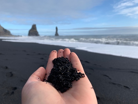 Black sand on the white female hand close up on the background of famous black sand beach Reynisfjara in Iceland and ocean landscape