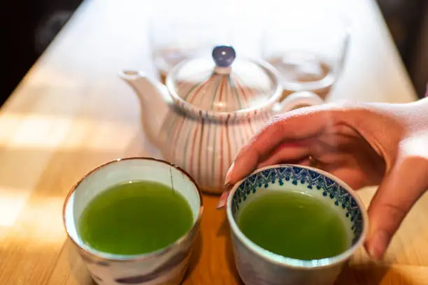 Woman hand closeup holding teacups or tea cups by teapot with hot traditional Japanese sencha green tea at kitchen wooden table