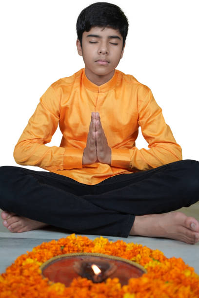 Vertical photo of one Indian Asian boy sitting boded hands and cross legged in front of a Pooja or prayer Thaali with a Jyot, Mitti ka Diya, or earthen lamp, Deepak flame and marigold flowers. The model is Rishabh Syal who is a pre teen boy from India, Asia. Apt for Diwali, Deepawali, Durga pooja, Rakshabandhan, Raakhi, Ganesh Chaturthi, Lakshmi Poojan festival, festivals. The boy is in an orange colored kurta and black jeans. There is a puja Aarti plate or thali with a jyot or lau, and yellow orange color auspicious gainde gainda ke phool, flowers. The child is in the centre, middle, center of the frame. White wall in the background no text, copy space. Front view pose, In prayer pose, eyes closed. fotografía vertical de un niño asiático indio, aislado sobre blanco, sentado con las piernas cruzadas en el suelo, las manos dobladas en namaste y los ojos cerrados, rezando con un pooja thaali con un jyot, de guantes ka diya, deepak o llama de lámpara
