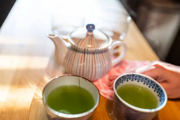 Woman hand closeup wiping wooden table with cloth or tablecloth by teapot with hot traditional Japanese sencha green tea cup in kitchen