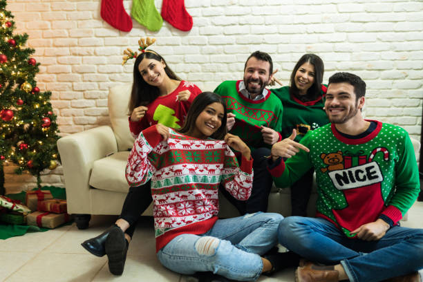 Friends In Ugly Sweater Celebrating Christmas Together At Home Cheerful friends showing off their ugly sweater while enjoying Christmas party at home christmas sweater stock pictures, royalty-free photos & images