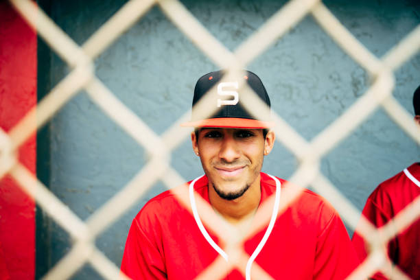 Relaxed and smiling Hispanic baseball player photographed through fence while sitting in dugout with teammates.