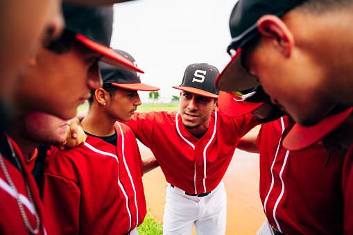 Interior viewpoint of young Hispanic baseball players huddled with arms around each other and listening to motivational talk from captain.