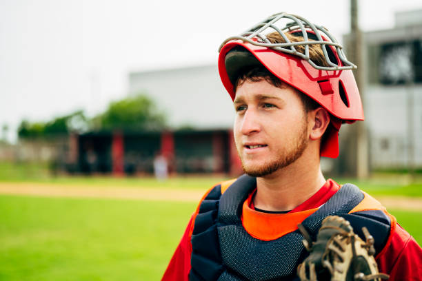 Young baseball catcher wearing mask and chest protector Outdoor portrait of young Hispanic baseball catcher with glove wearing chest protector and mask and looking away from camera. catchers mask stock pictures, royalty-free photos & images