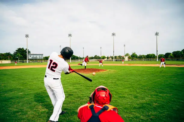 Rear viewpoint of Hispanic baseball player standing in batter’s box and swinging his bat at thrown pitch.