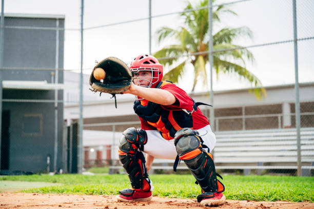 Baseball catcher crouching to catch pitch in center of mitt Low angle action portrait of baseball catcher wearing helmet, chest protector, and leg guards crouching with mitt poised to catch pitch. high school baseball stock pictures, royalty-free photos & images