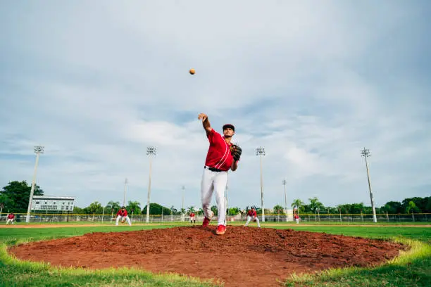 Low angle view of Hispanic baseball player delivering a pitch from the mound with alert infielders in the background.