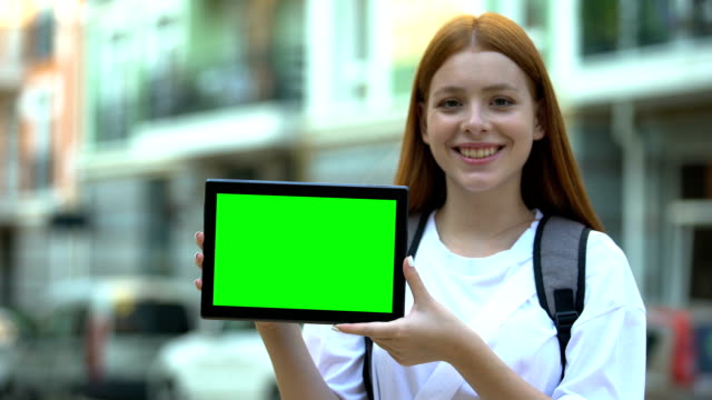 Smiling girl with backpack showing green screen tablet, cheap tours and flights