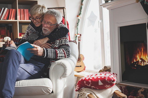 Shot of senior married couple enjoying a cold winter day while sitting next to a fire place in their living room and cuddling while reading a book.