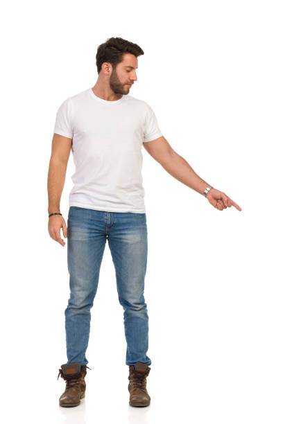 istock Serious Handsome Man In Jeans And White T-shirt Is Standing And Pointing Down. Front View. 1174902708