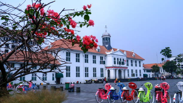 Early morning traffic of motorcycles and cars passing by a row of colonial-era buildings along Jalan Braga in Bandung, Indonesia. The sunlight casts a soft haze over the scene, highlighting both the vibrancy of urban movement and the charm of preserved Dutch architecture.
