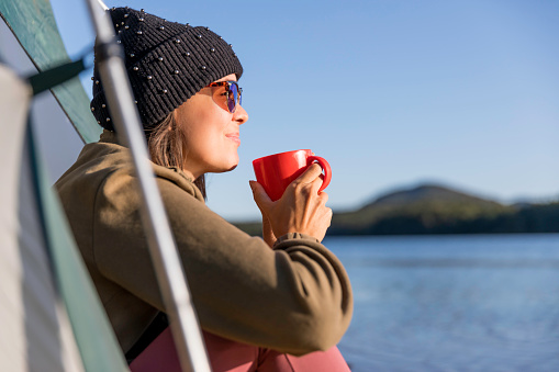 A young women is drinking a coffee by the lake in camping. She is sitting in her tent and contemplating the view. She is wearing warm clothing.