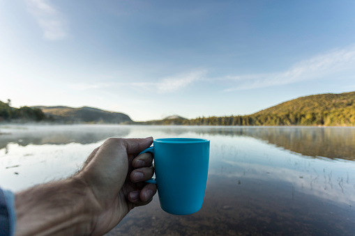 A man is drinking a coffee by the lake in camping in Autumn. He is sitting on a chair and contemplating the view. You can see his personal perspective of the nature surrounding him.