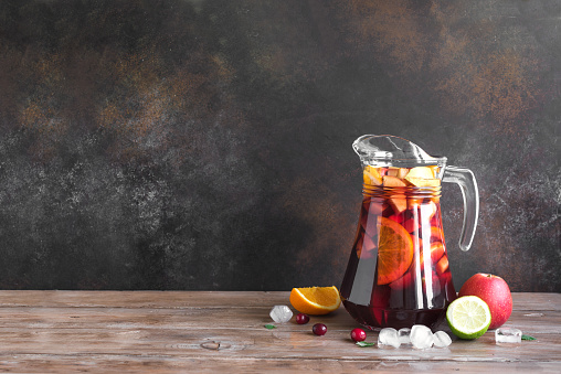 Red wine sangria or punch with fruits and ice in pitcher. Homemade refreshing fruit sangria on wooden background, copy space.