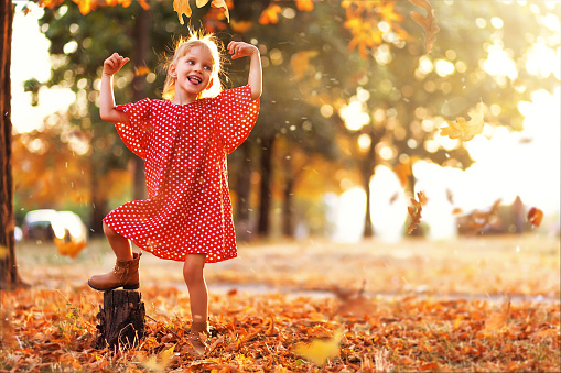Journey in nature. Adorable happy girl throwing the fallen leaves up, playing in the autumn park. Little blonde hair and blue eyes girl smiling expression. Girl in polka dot red dress and boots