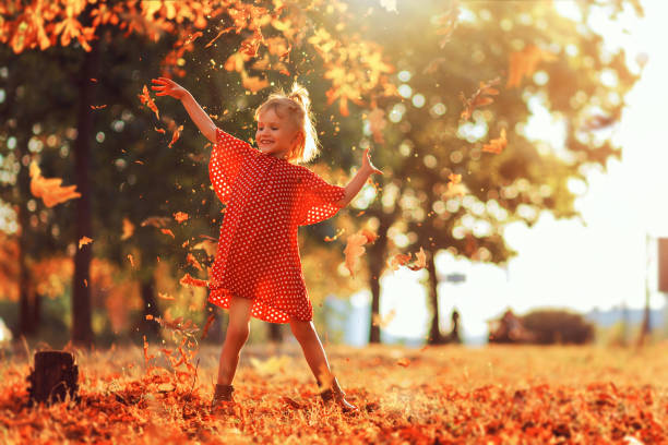 viagem na natureza. menina feliz adorável que joga as folhas caídas acima, jogando no parque do outono. cabelo louro pequeno e expressão de sorriso da menina dos olhos azuis. menina no vestido e em carregadores vermelhos do ponto de polca - little girls autumn child red hair - fotografias e filmes do acervo