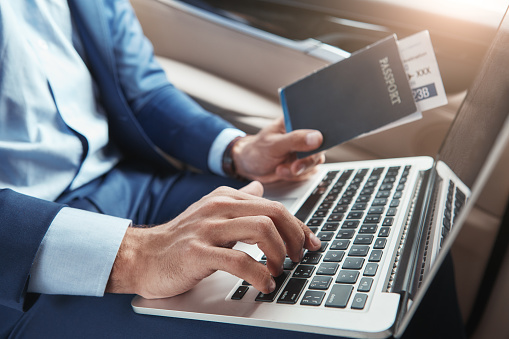 Check in. Cropped image of young businessman in formal wear working on laptop and holding passport with flight tickets while sitting in car. Travel concept. Business concept. Digital technologies