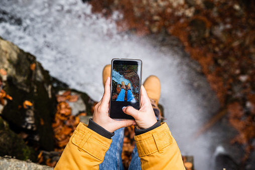 woman taking picture on phone of her legs with waterfall on background autumn season