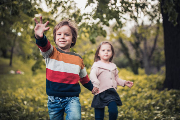 Two happy kids having fun while holding hands and running in spring day at the park. Focus is on boy waving.