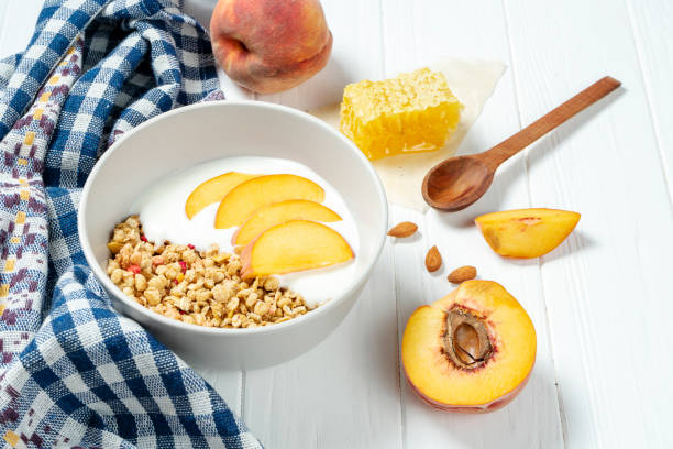 istock white wheat granola with low-fat yogurt in a white bowl in a composition with a spoon, honeycombs and peach on white wooden background. Healthy breakfast food. Making breakfast. Gluten free 1174939157