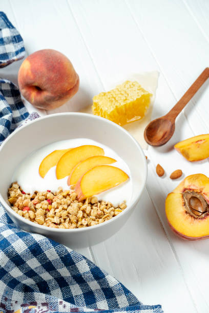 istock white wheat granola with low-fat yogurt in a white bowl in a composition with a spoon, honeycombs and peach on white wooden background. Healthy breakfast food. Making breakfast. Gluten free 1174939173