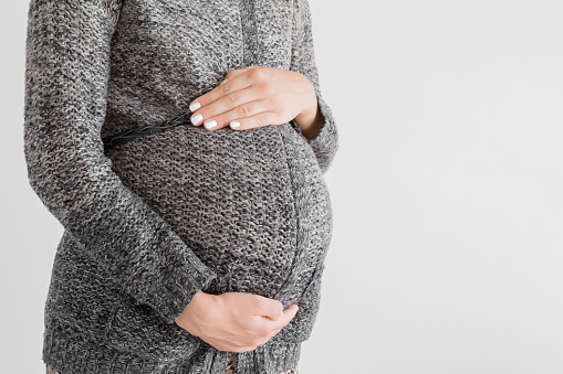 Pregnant woman in knitted dark jacket touching big belly with hands. Isolated on gray background. Warm clothes in cold weather in pregnancy time. Baby expectation. Side view. Closeup.